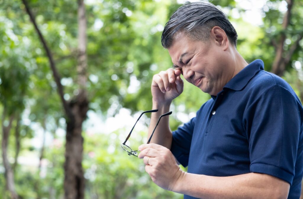 A man standing outside in a park holding his glasses in his left hand as he rubs his right eyes with his right hand.