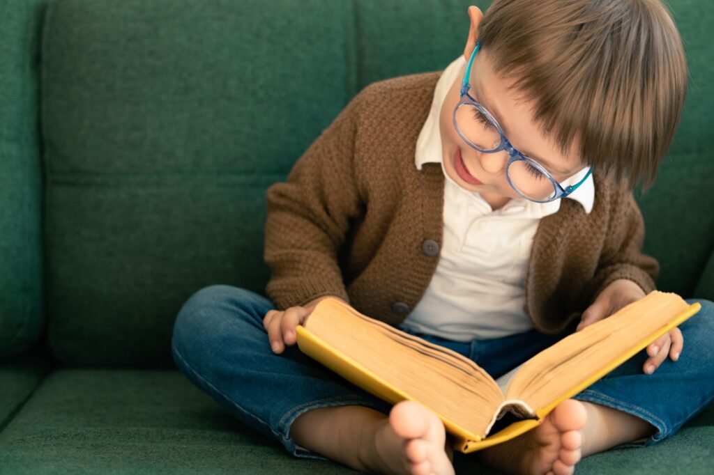 Barefoot toddler wearing glasses reads a large book on a green couch.