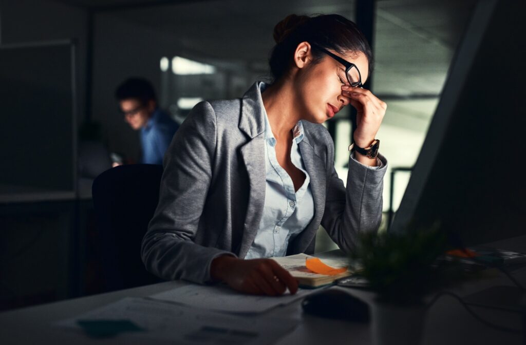 A person sits in front of a computer, eyes closed and fingers pinching the bridge of their nose, visibly uncomfortable from dry eye irritation.
