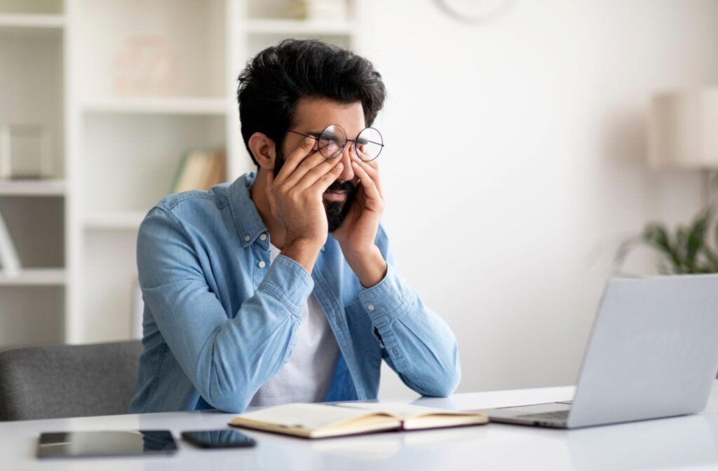 Man sitting at a computer with eyes closed tightly, hands rubbing eyes, experiencing digital eye strain.
