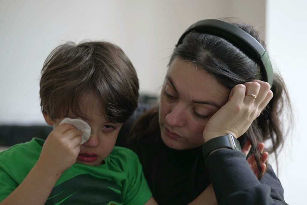 A young boy rubbing his irritated eye with a cotton pad while sitting next to a concerned woman who appears to be comforting him.