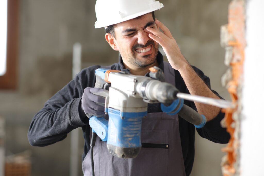 A construction worker in a hard hat wincing and rubbing his eye while holding a power drill, likely injured by flying debris.