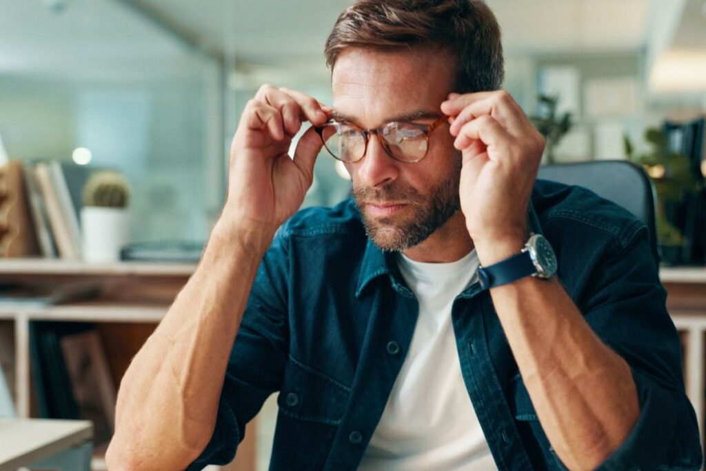 Adult man holding his glasses on his face as he works on a computer.